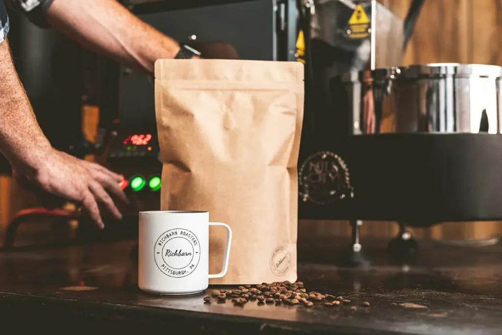 Sachet kraft, tasse et grains de café disposés devant un torréfacteur.